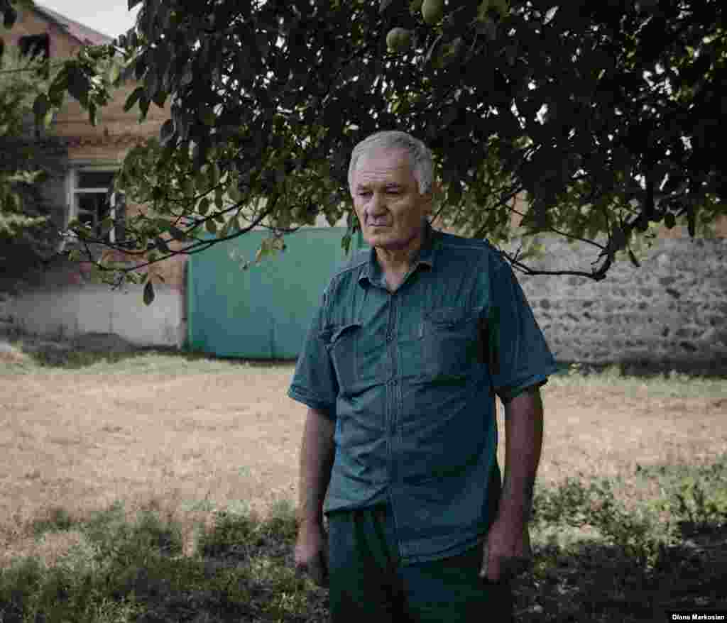 «Мені здається, що вона як і раніше з нами», – говорить 63-річний Руслан Коков. Коли Оксана пішла в школу, щоб більше вже не повернутися ніколи, Руслан був на базарі. В її день народження Руслан і його 27-річний син Урузмага завжди сервірують для неї місце за столом. Вони їдять її улюблену їжу – кавуни, хурму, апельсини-корольки. На цій фотографії Руслан зображений на тлі свого будинку на вулиці Комінтерну. Школу №1 видно з вікна його кімнати. Під час облоги тут розташовувалися підрозділи російського спецназу. У заключній перестрілці було розбито скло на двох вікнах його будинку. За минулі десять років він так і не зібрався з духом їх полагодити. «Коли щось нагадує мені про цю трагедію, я тільки страшно засмучуюсь», – говорить він. 